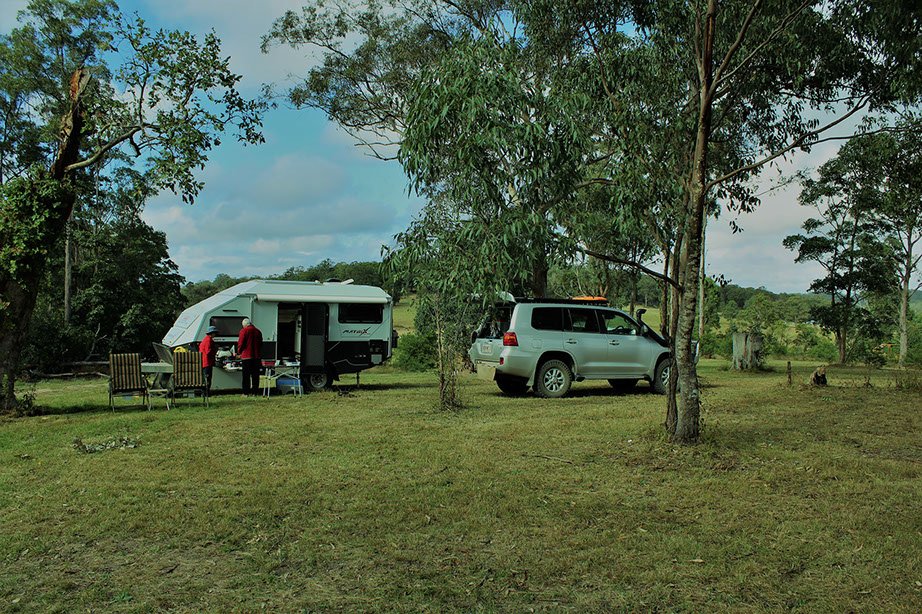 A camping scene with a white camper trailer, a silver SUV, and two people in red jackets standing outside near a table and chairs in a grassy area surrounded by trees.