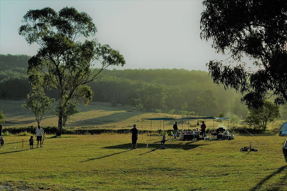 People standing and sitting under a canopy in a grassy field with trees in the background, possibly at a park or outdoor gathering during late afternoon or early evening.