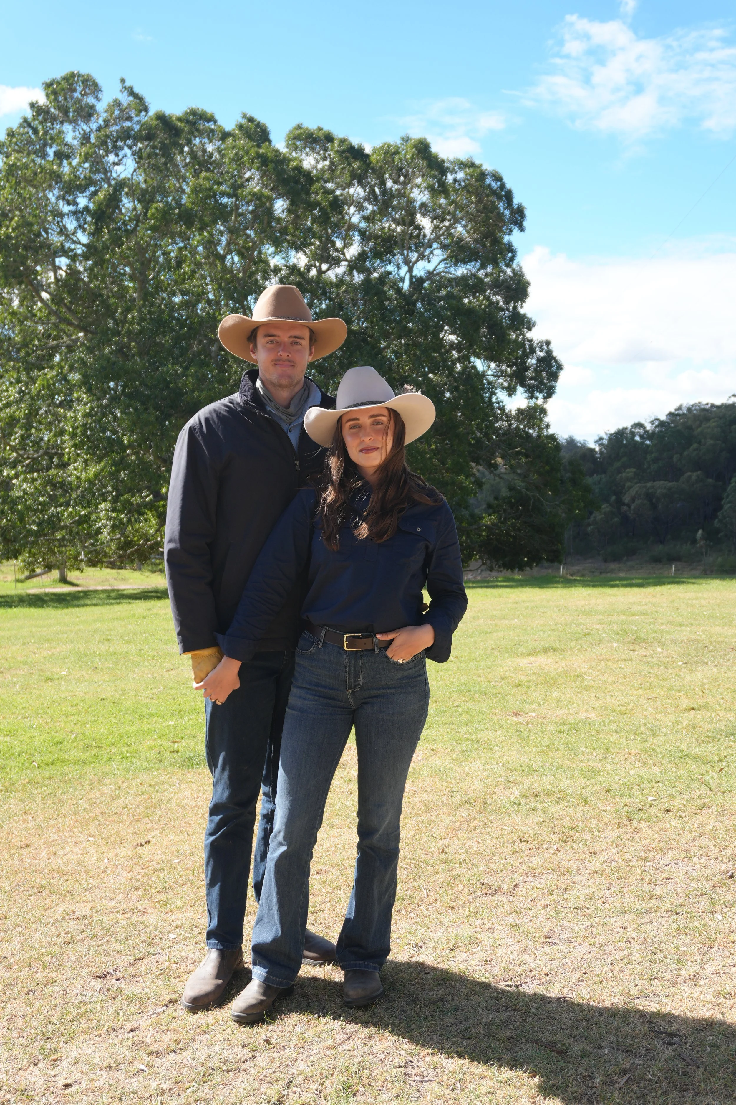 A man and woman standing outdoors with trees and a blue sky in the background. They are wearing cowboy hats and casual clothing.