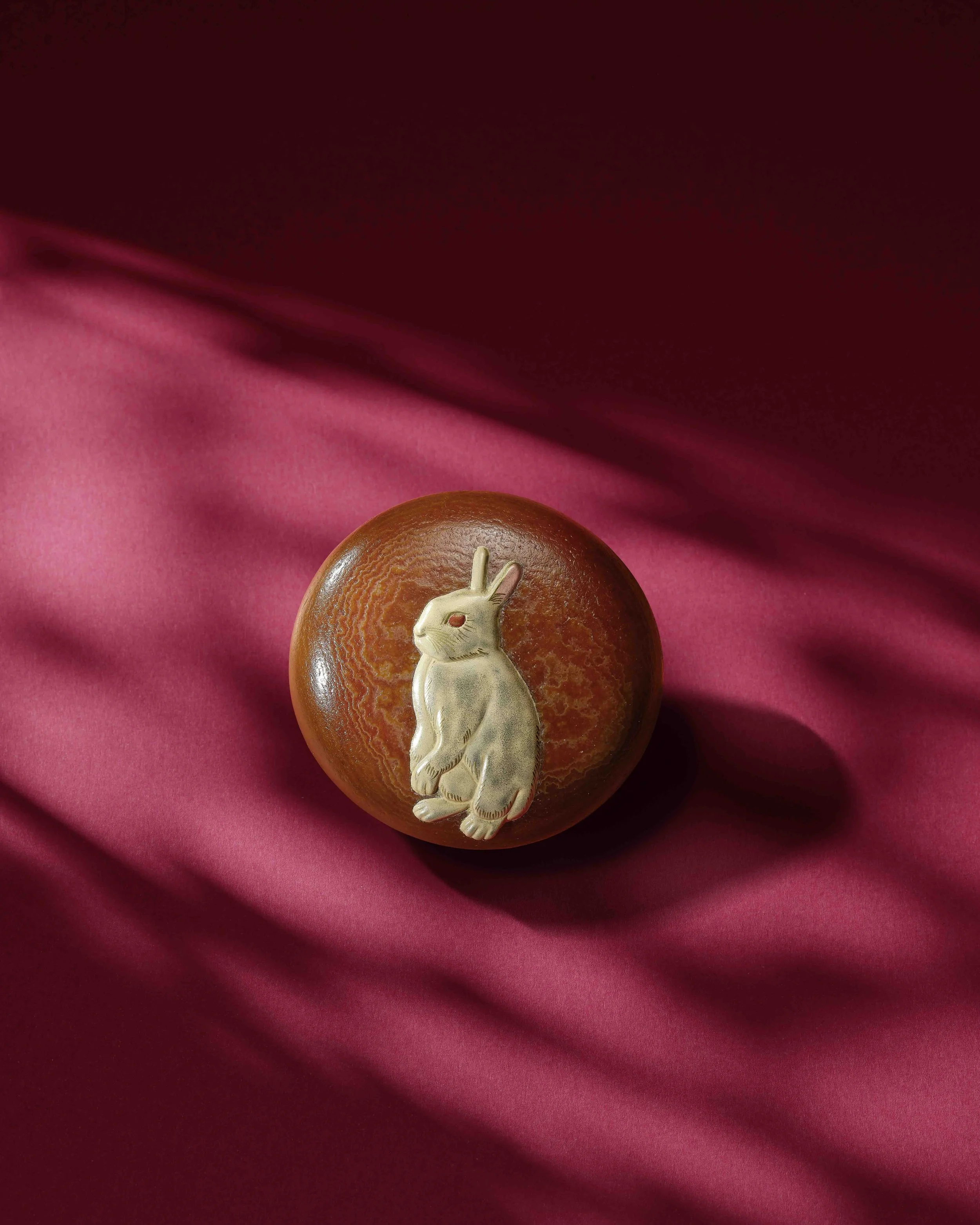 A mottled russet and golden agate pebble of circular form, worked in silver hiramakie and takamakie with a seated rabbit, the detail of the eye in red lacquer, 6 x 6 cm