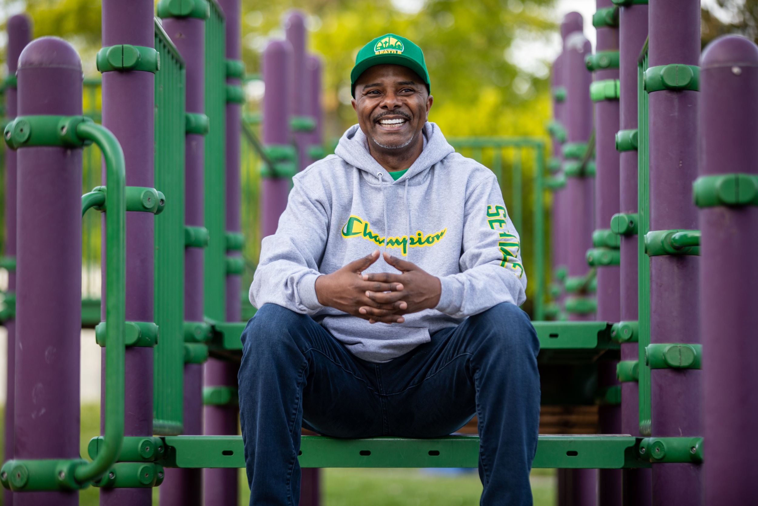 Photo of a man wearing a sweatshirt that reads "Champion" and on his sleeve "Seattle" seated in a purple and green playground looking off into the distance.