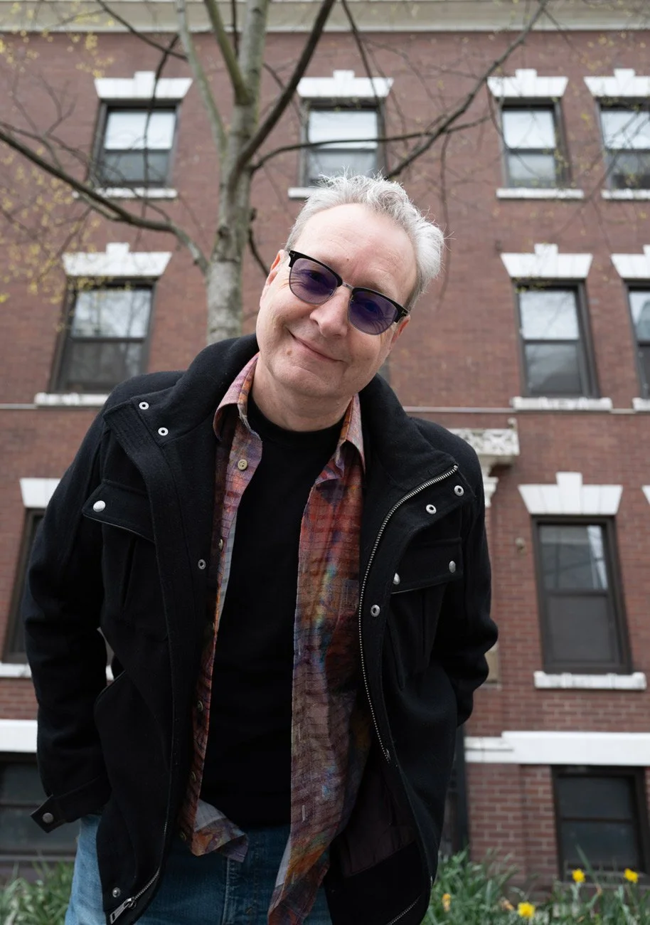 Photo of a man looking off to his left wearing a black jacket and blue jeans with sunglasses on. In the background is a brick wall and two windows as well as a lamp.