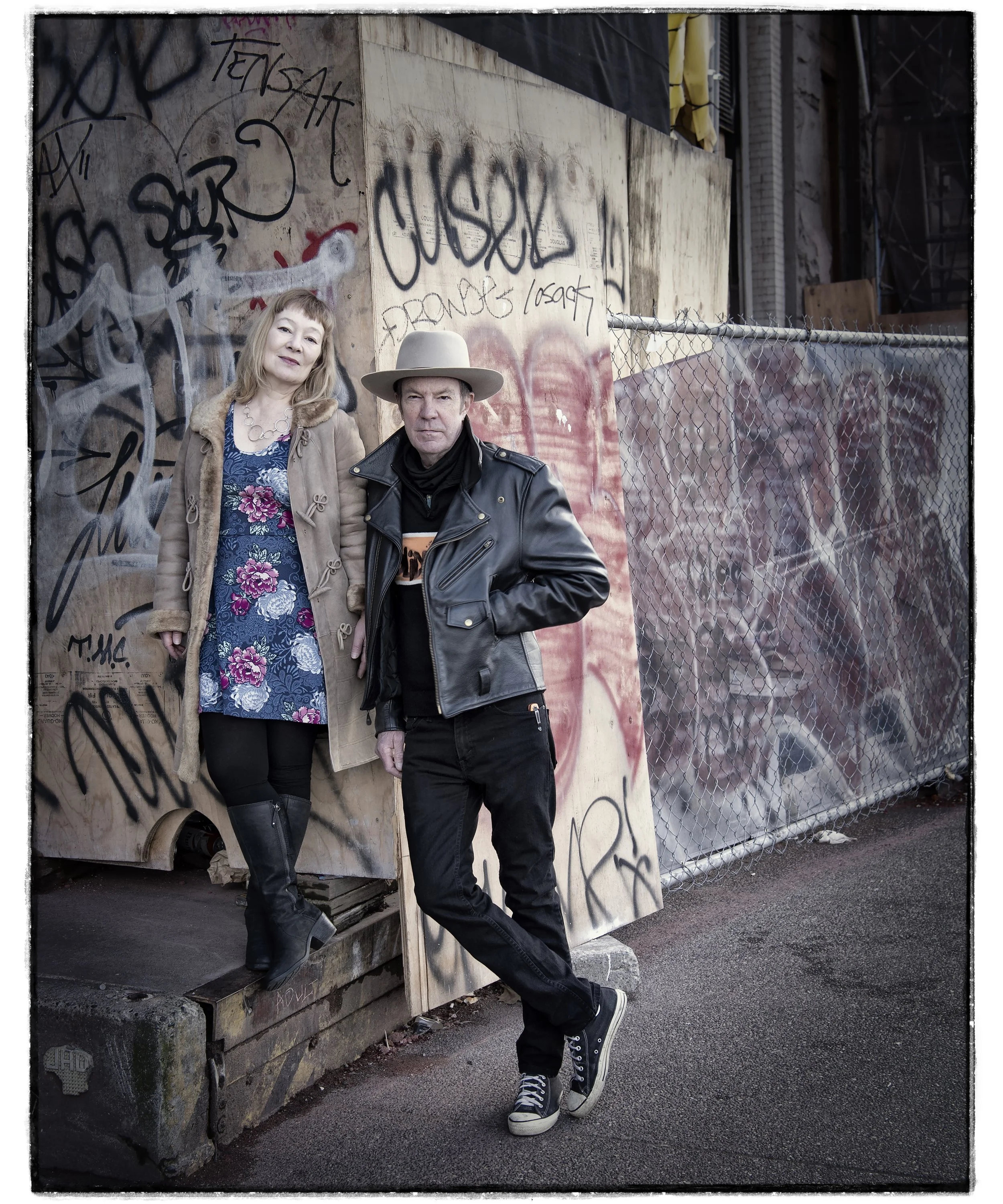 Photo of a man and woman in front of a construction site. The man is wearing a leather jacket and black jeans with a large  hat. The woman is wearing a brown jacket, a blue dress with flowers, and wearing black leather boots.