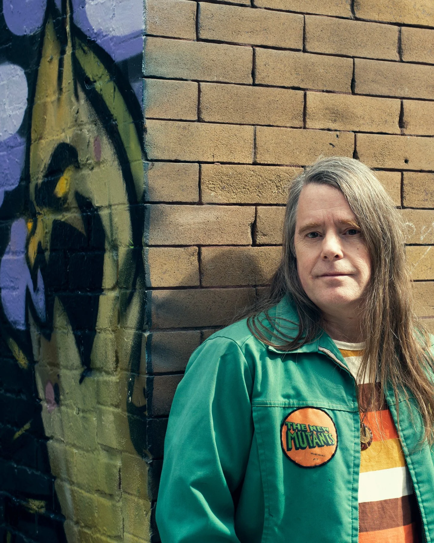 A photo of a man with long hair wearing a green jacket with a patch that reads "The New Mutants" against the corner of a brick wall with a mural on one side.