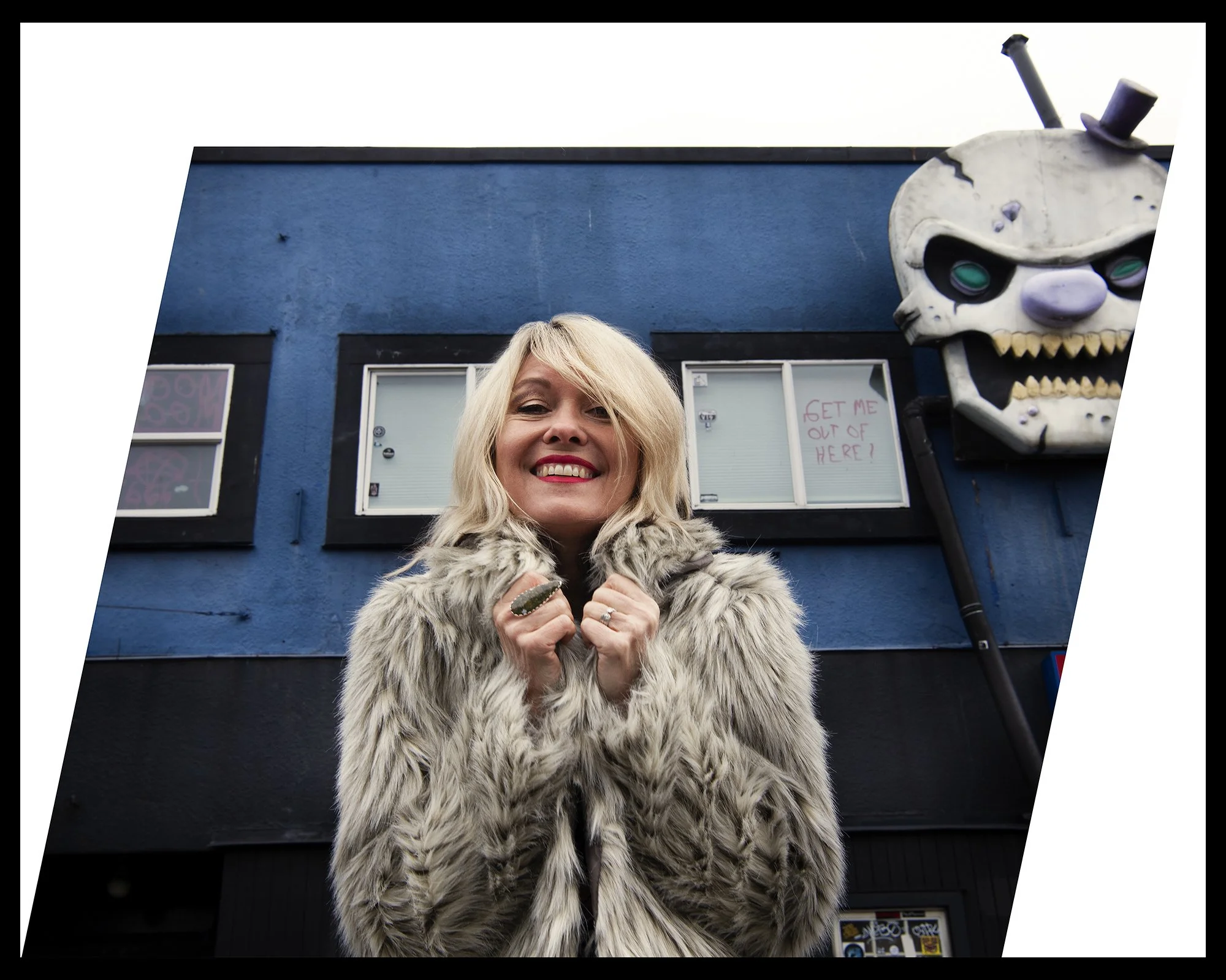 Photo of a woman with long blond hair and a fur coat. In the background is a blue building with a large skull.