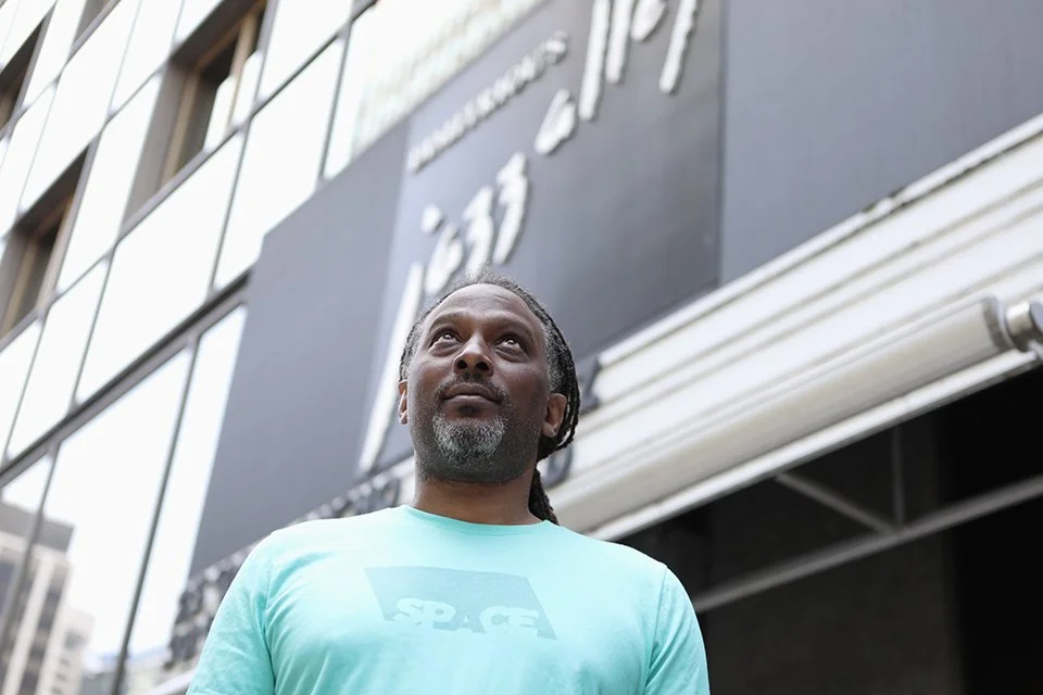 Photo of a man in a blue t-shirt looking up. In the background behind the man's head reads Dimitriou's Jazz Alley in large letters.