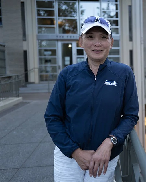 Photo of a woman wearing a hat with sunglasses on her head and a blue jacket with the Seahawks logo in white pants. She is standing in front of the Exhibition Hall at the Seattle Center.