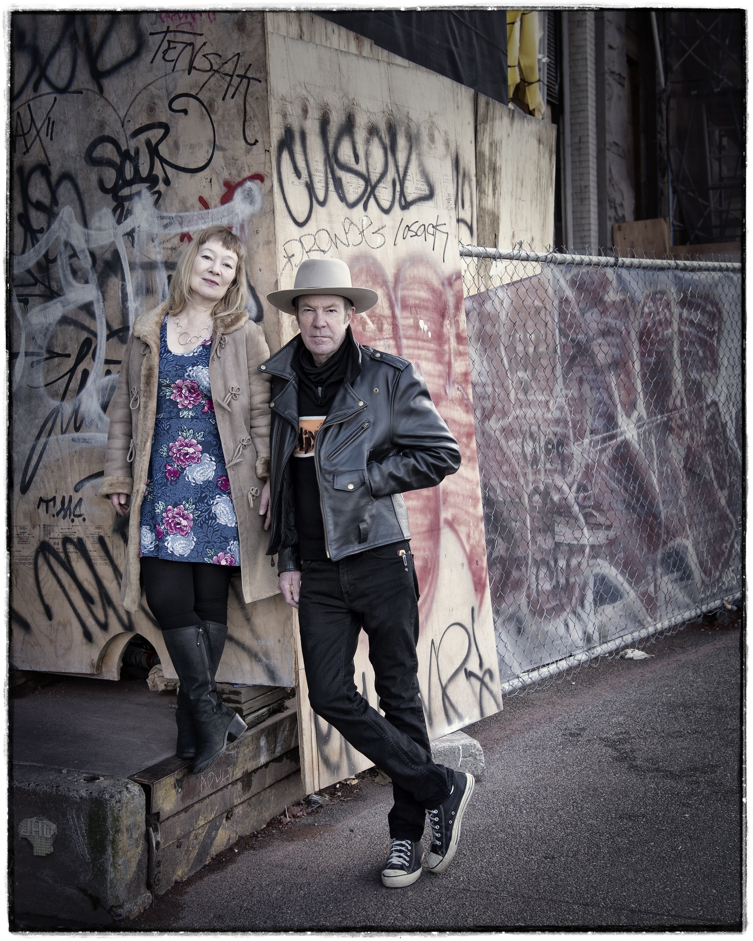 Photo of a man and woman in front of a construction site. The man is wearing a leather jacket and black jeans with a large  hat. The woman is wearing a brown jacket, a blue dress with flowers, and wearing black leather boots.