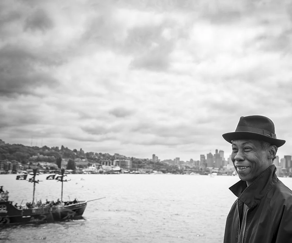 Photo of a man with a black fedora and jacket looking off to the left of the frame in blakc and white. In the background is the skyline of Seattle.