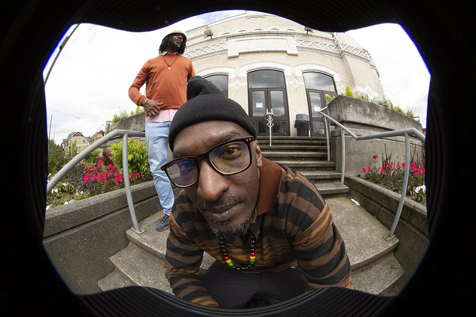 Photos of two men, one seatted wearing a black and brown collar shirt with glasses and a neckless and another man standing in the background wearing an orange sweater and blue jeans in front of a high school.