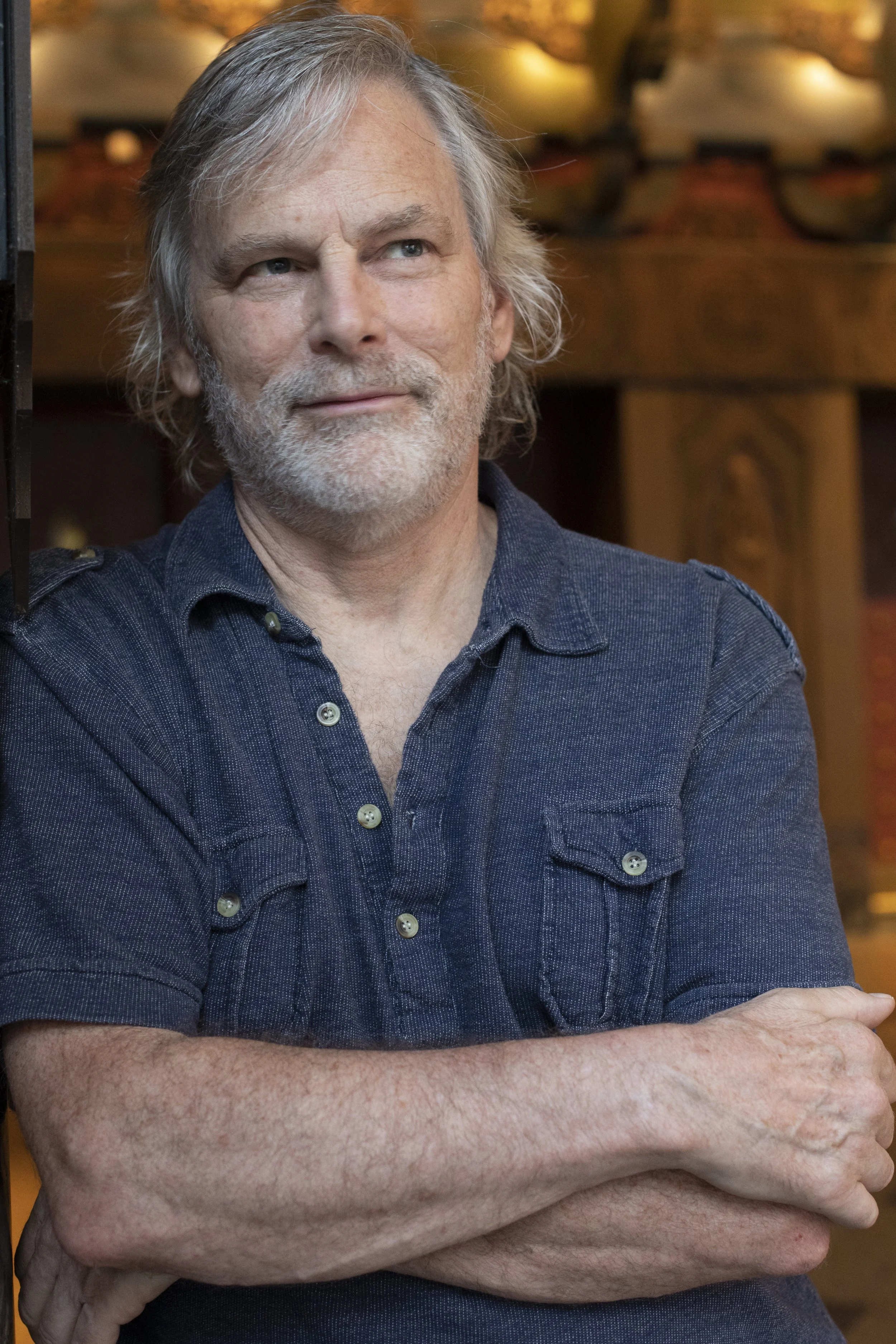 Photo of a man with medium length white hair and a blue button-down shirt looking off into the distance. Blurred in the background is the backdrop of a theater facade.