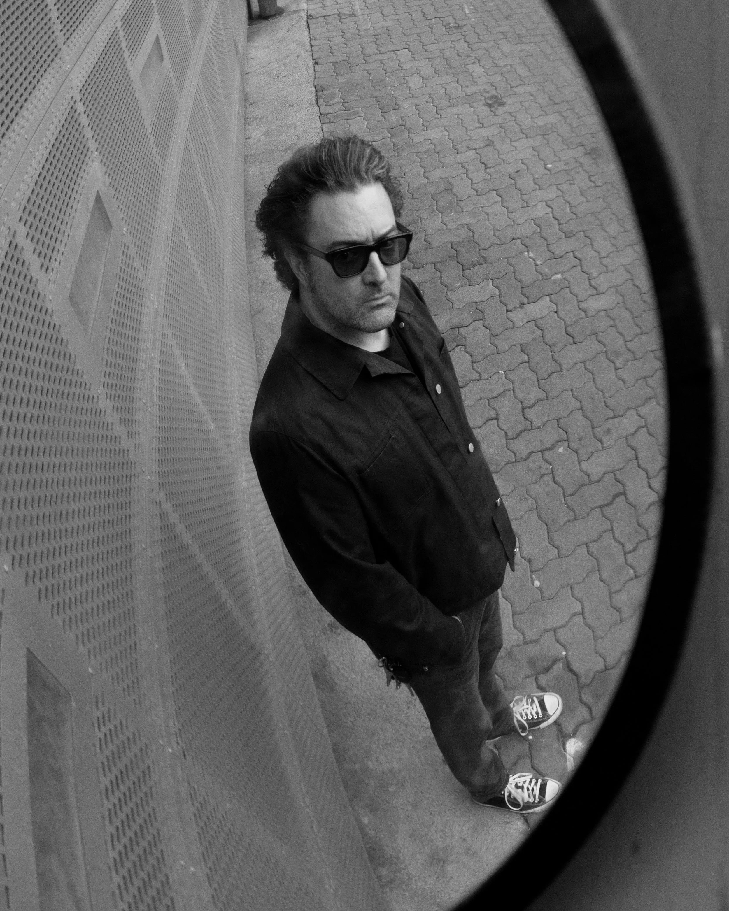 photo of a man wearing sunglasses with a black shirt and jeans peering into a distorted mirror. Photo is black and white.