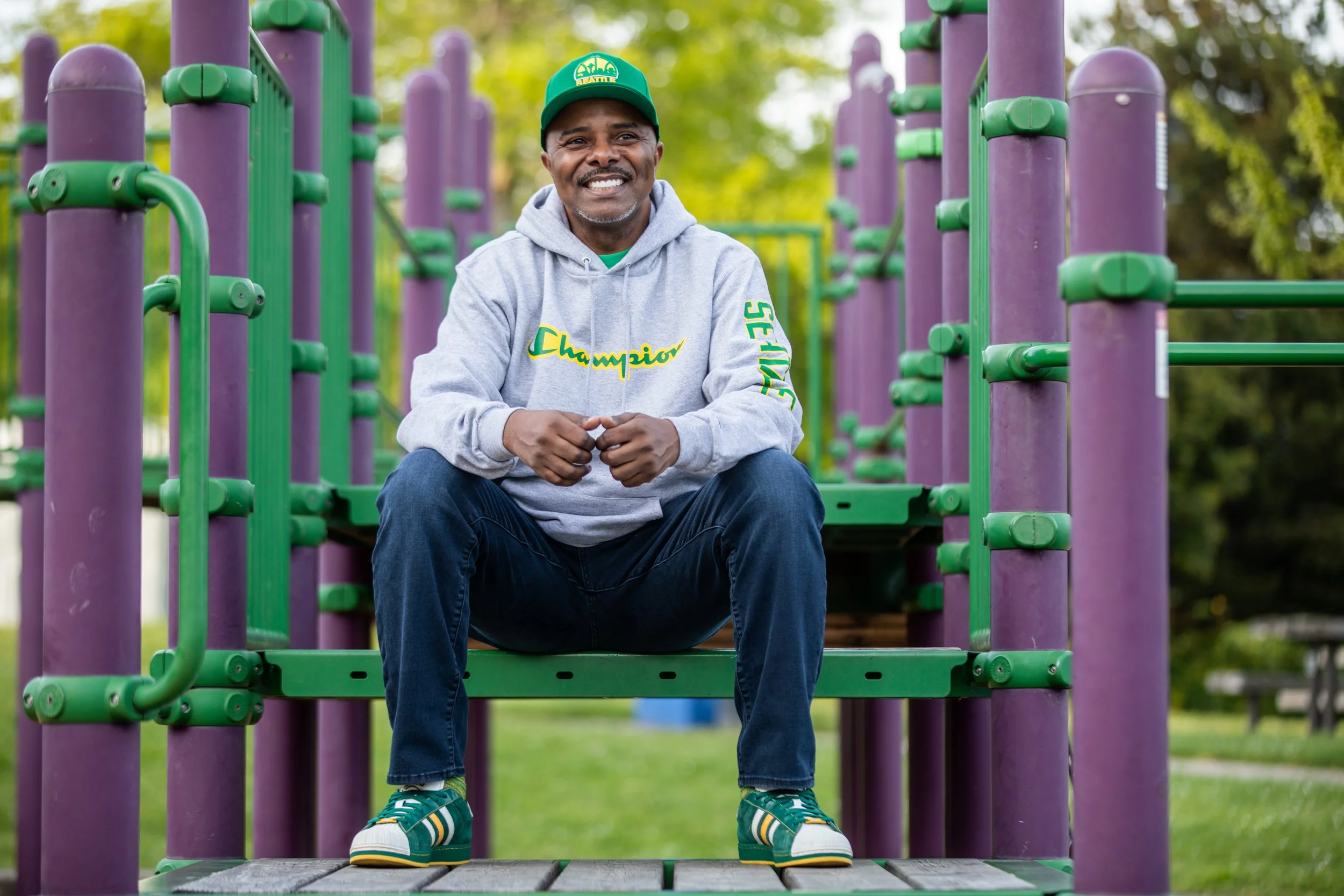 Photo of a man wearing a sweatshirt that reads "Champion" and on his sleeve "Seattle" seated in a purple and green playground looking off into the distance.