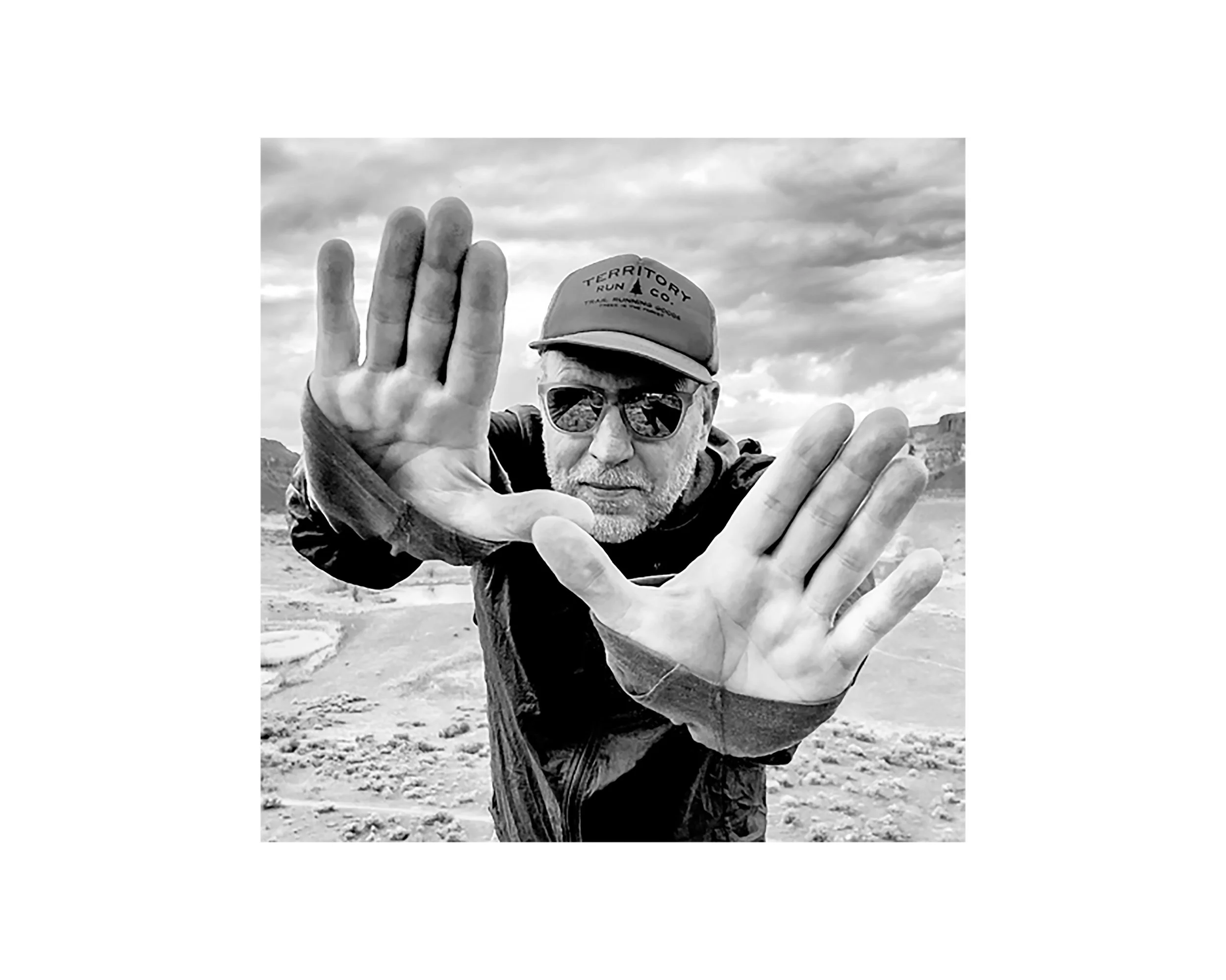 black and white photo of a man with his hands up wearing a hat that reads "Territory Run Co." and sunglasses.