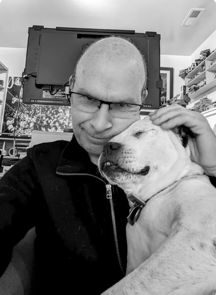 black and white photo of a man wearing a black jacket holding a white dog under his left hand.