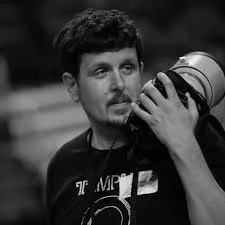 Photo of a man wearing a t-shirt and a large camera lens over his shoulder.