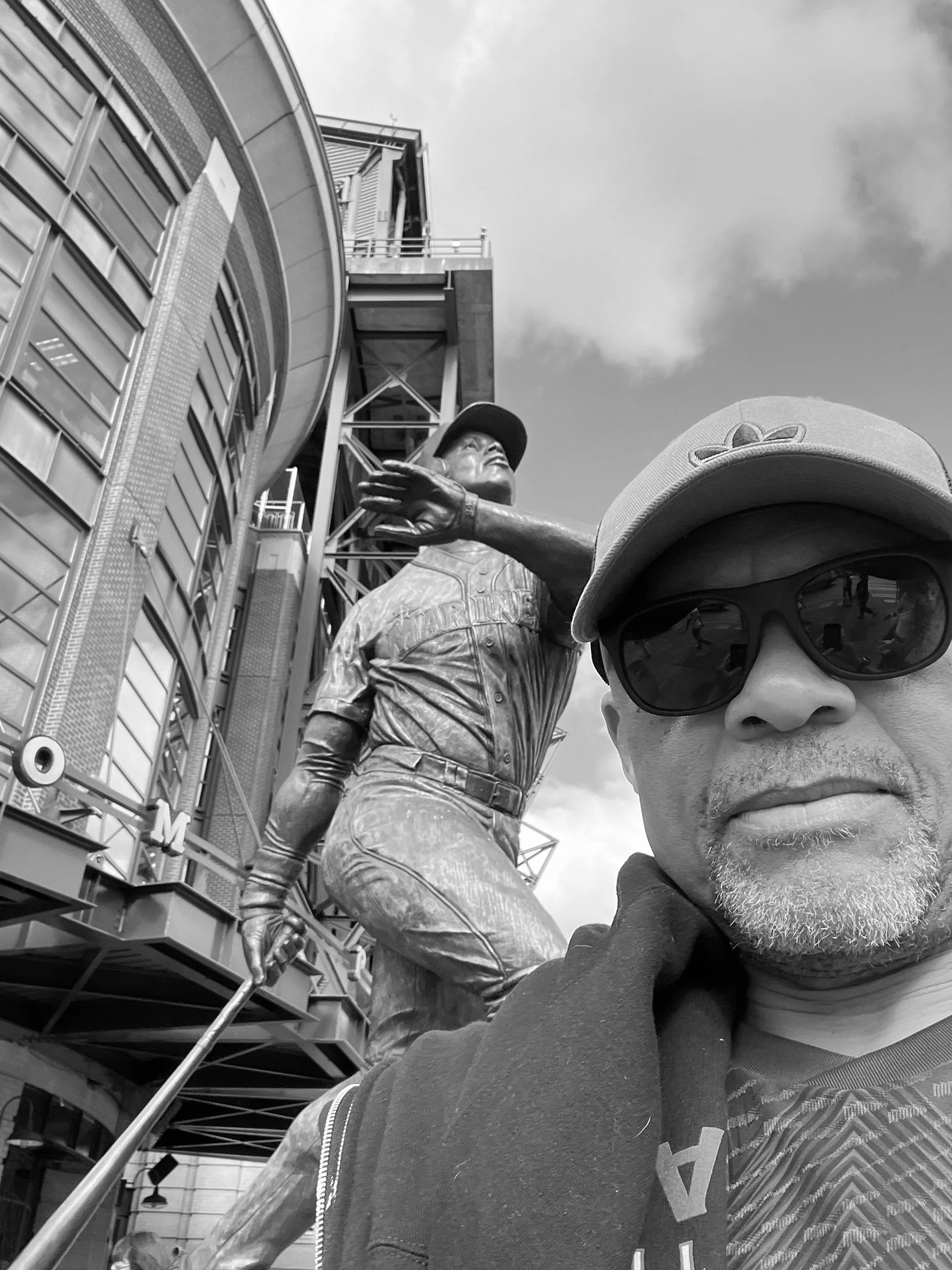 Photo of a man with a hat and sunglasses on in front of a statute of a baseball player.