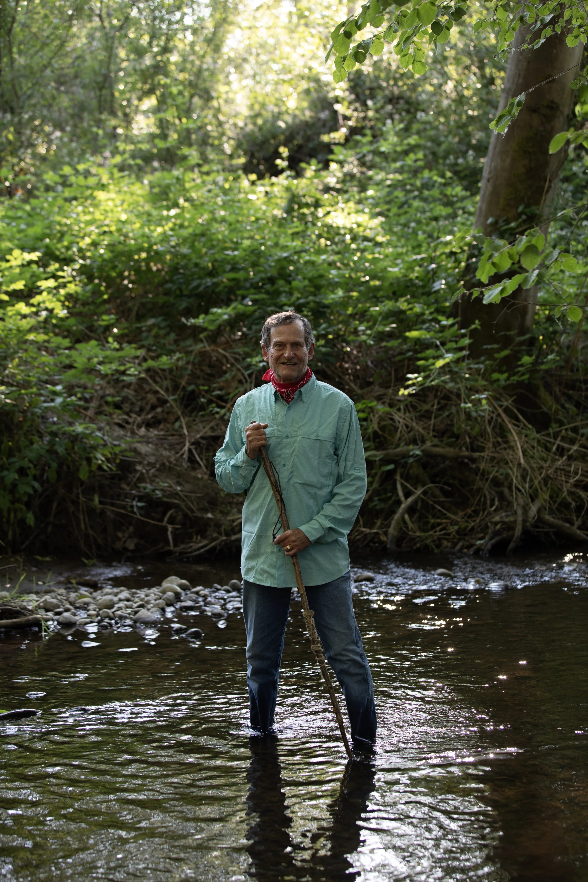 photo of a man in a green shirt and blue jeans standing in a creek surrounded by nature.