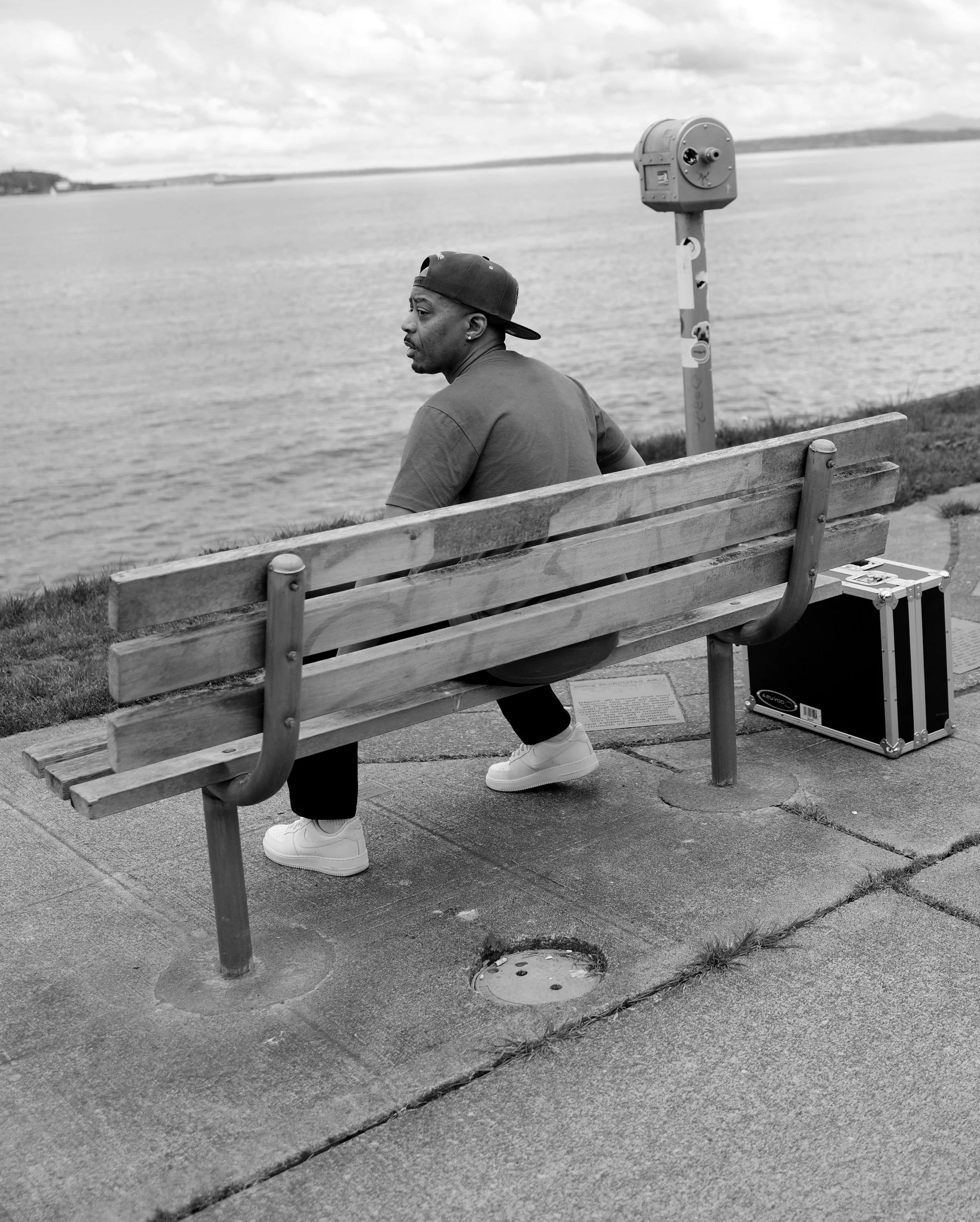 Photo of a man wearing a dark t-shirt sitting on a park bench facing away from the camera in black and white. In front of him is the Puget Sound.