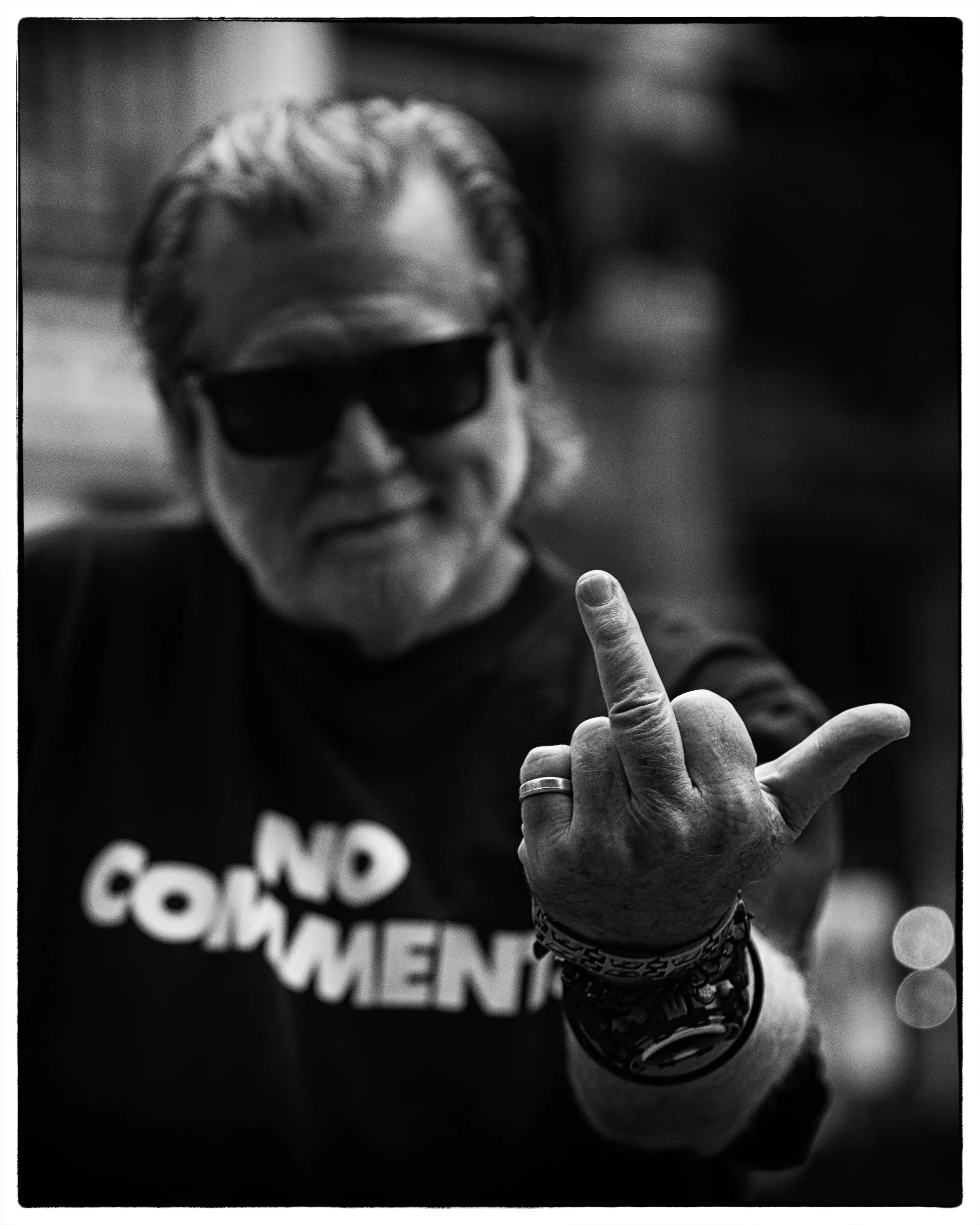 Photo of man in black and white with a white beard, sunglass on, and a t-shirt that reads "No Comment", blurred. In the foreground he is making a lewd gesture at the camera.