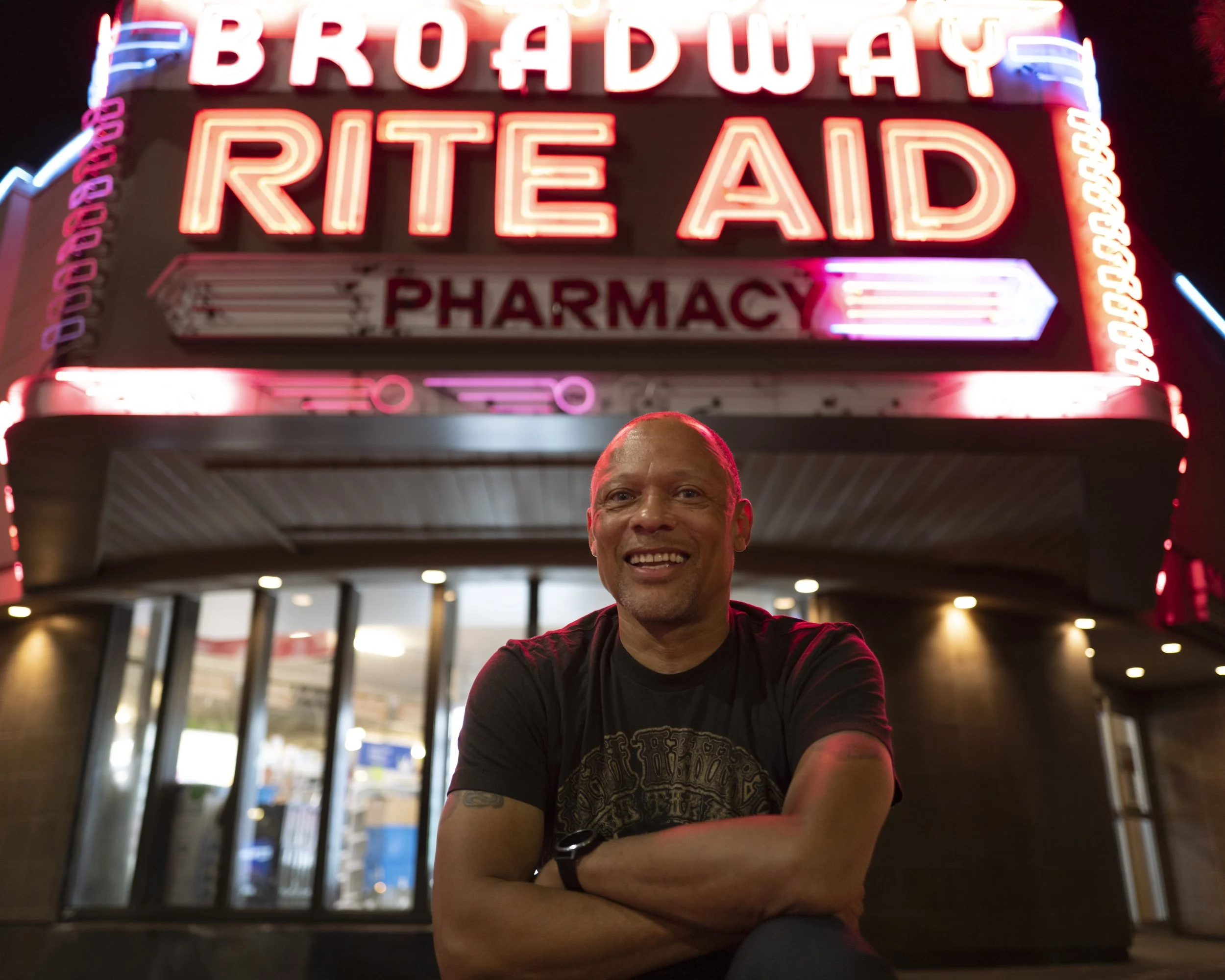 Photo of a man wearing a black t-shirt kneeling in front of a "Rite Aid Pharmacy" marque shown in neon.