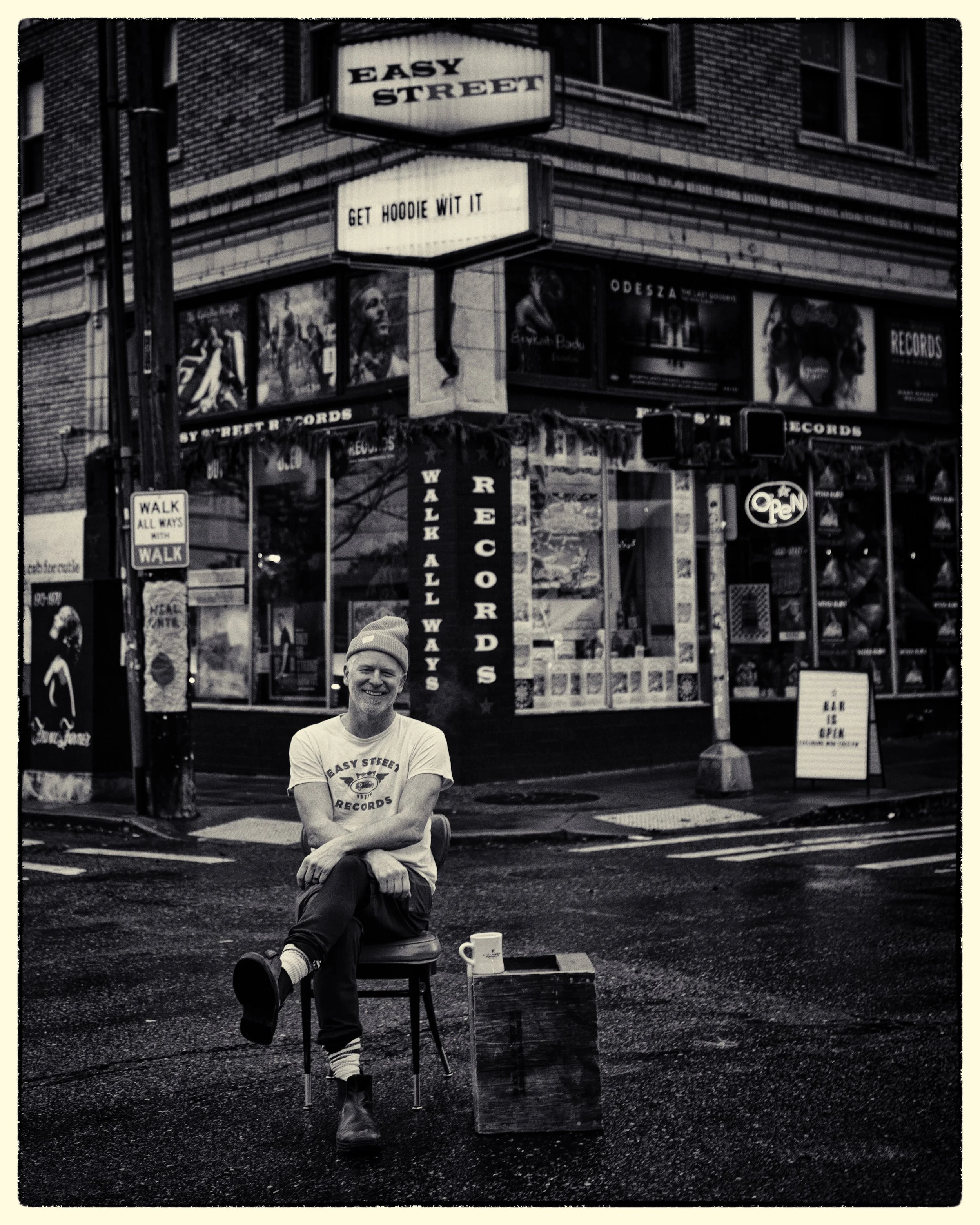 Photo of a man seated with a a beanie on, in the middle of the street in front a record store. Next to him is a wooden box with a coffee mug on top.