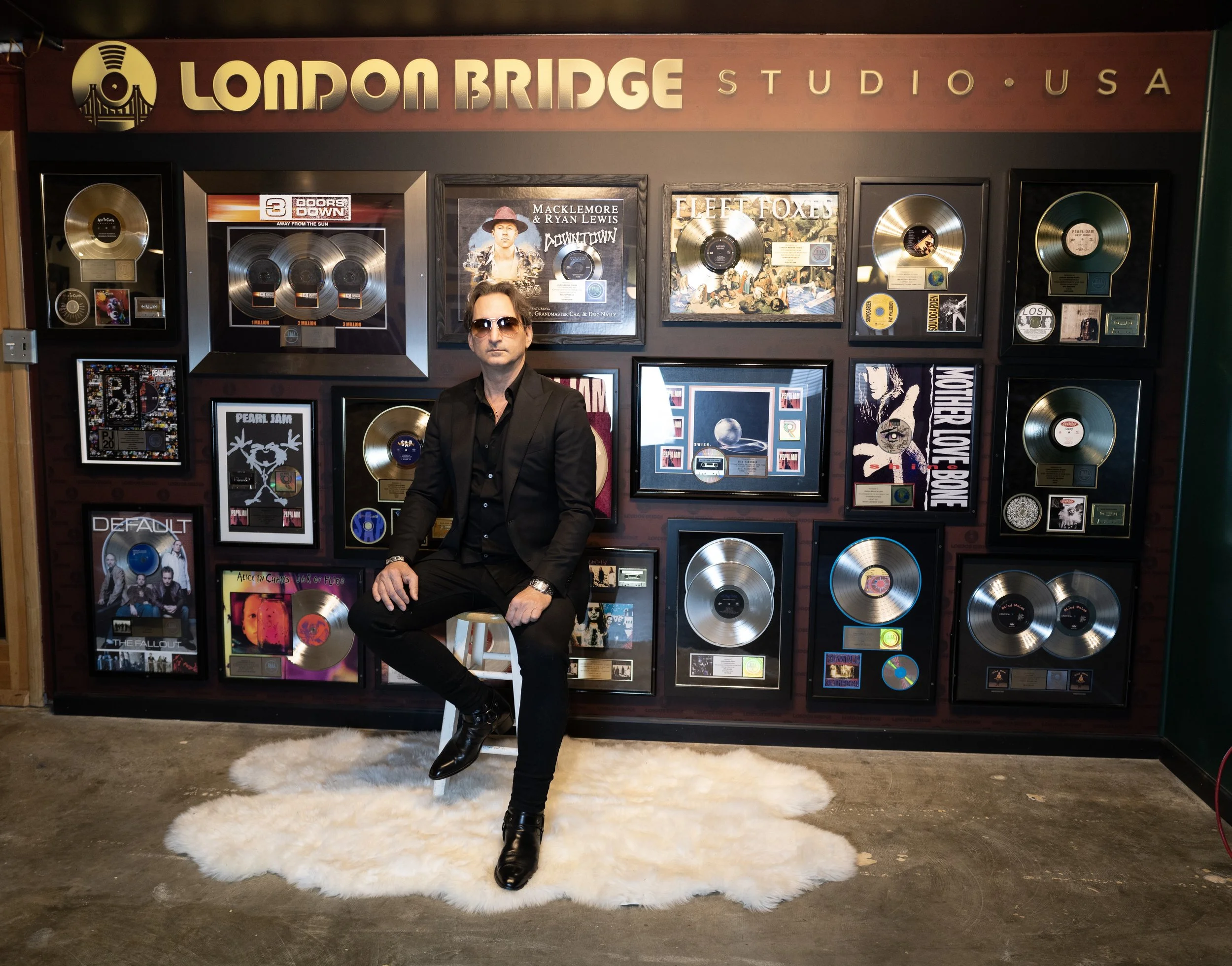 Photo of a man in a black sportscoat and sunglasses seated in front of a display of gold records. Above the records reads the words "London Bridge Studios".