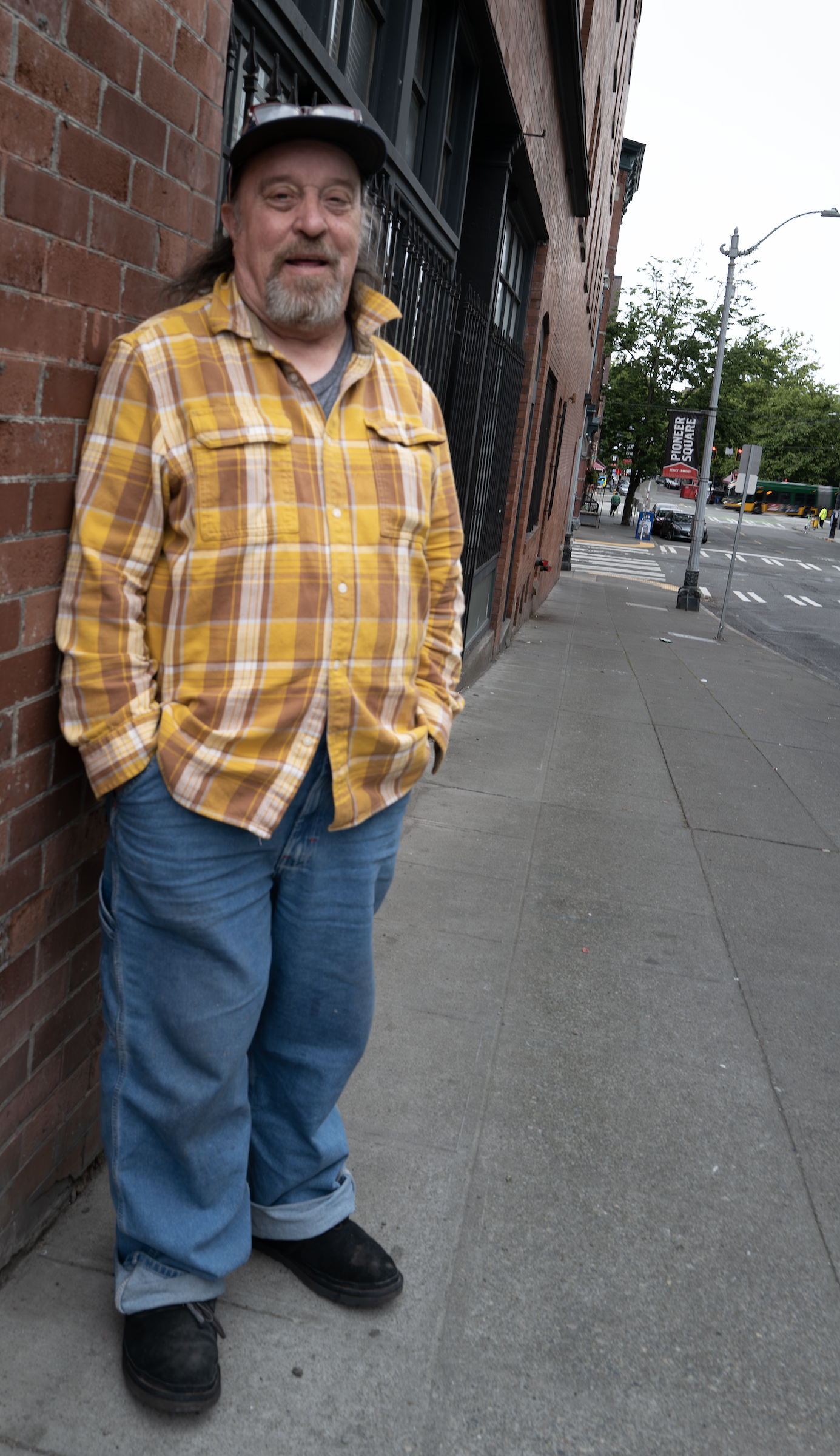 Photo of a man in a yellow, brown, and white patterned button-down shirt against a brick wall. In the background the words "Pioneer Square" can be read on a streetlamp.