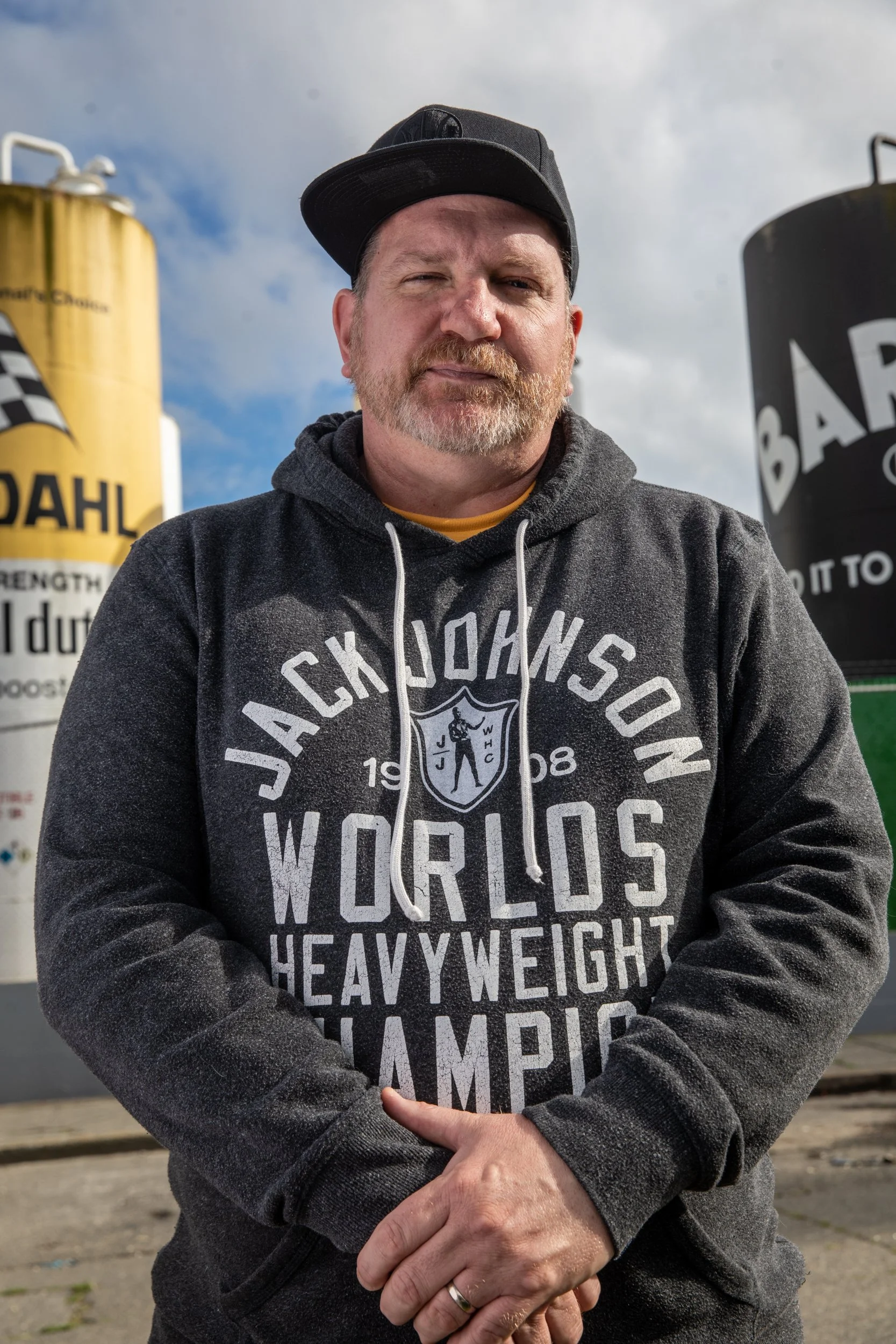 photo of a man in a black hat and grey sweatshirt with the words "Jack Johnson" standing in front of two large pieces of industrial equipment.