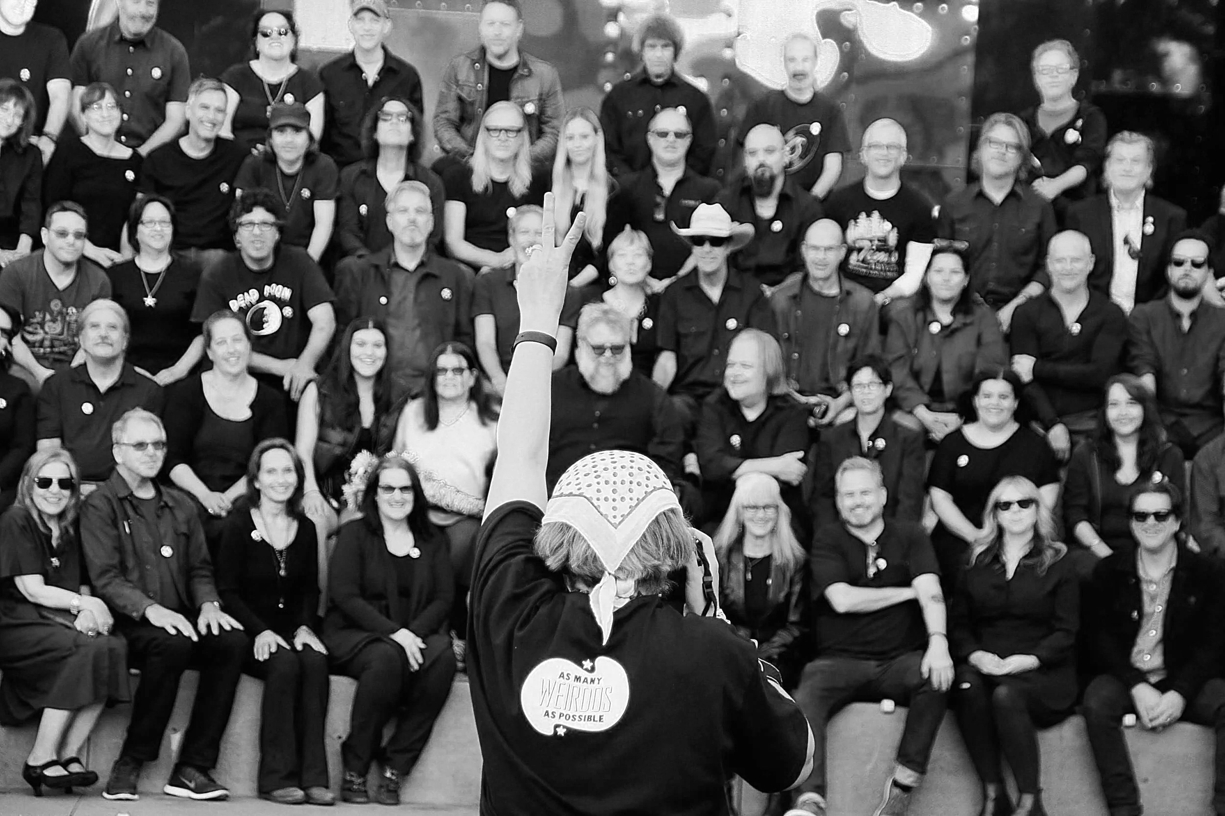 Black and white photo of a woman in a shirt with the text "as many weirdo as possible" on the black with a polkadot bandana on her head. the woman is holding a camera in her right had and making a peace sign with her left at a crowd of people sitting
