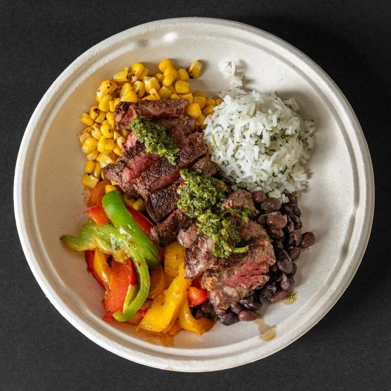 A bowl of food with white rice, grilled beef, black beans, corn, and mixed peppers topped with green sauce.
