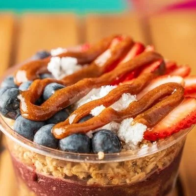 Close-up of a dessert in a glass with blueberries, strawberries, whipped cream, caramel drizzle, and granola topping.