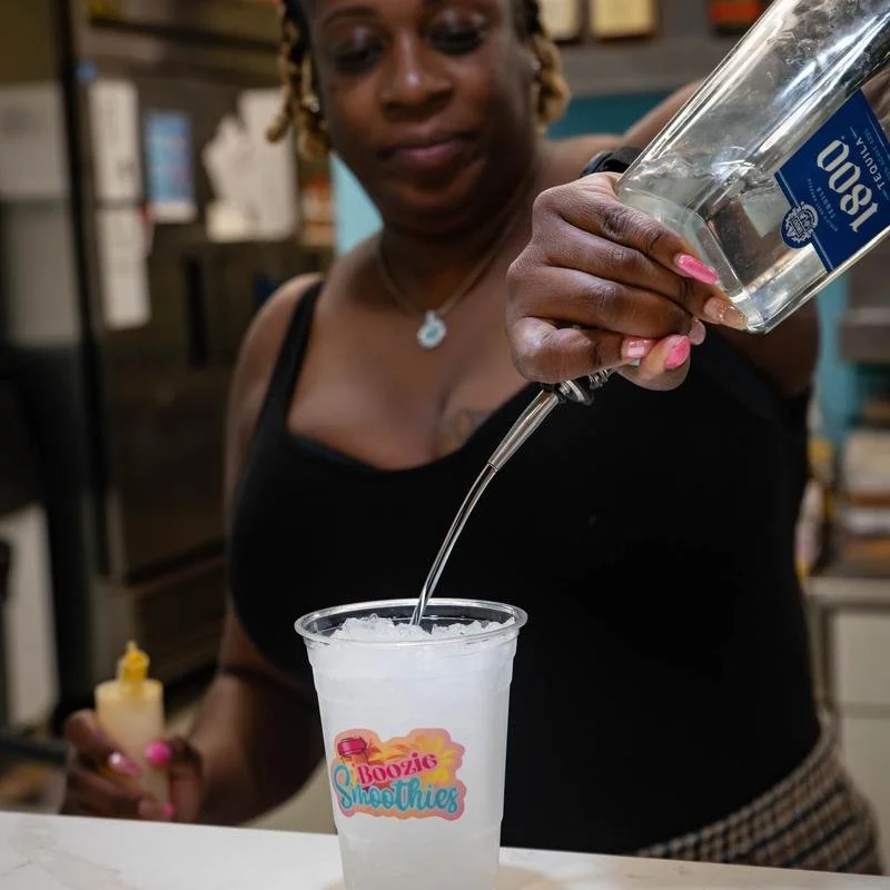 A woman with curly blonde hair and a necklace pours clear liquid from a bottle into a cup with a 'Boozie Smoothies' sticker on it.