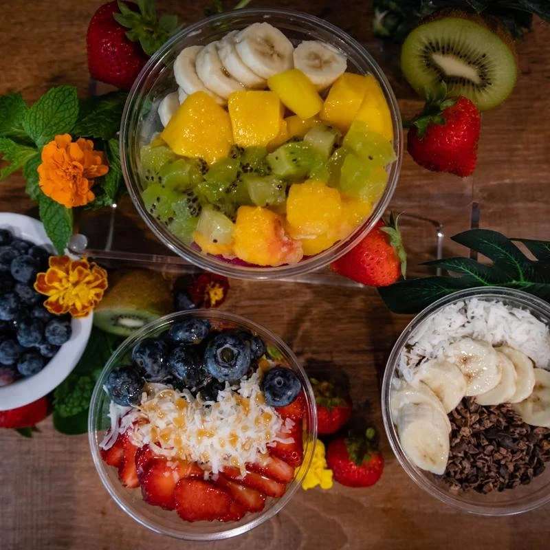 Three bowls of fruit parfaits topped with bananas, strawberries, blueberries, shredded coconut, and chocolate chips, with additional sliced bananas, kiwi, mango, strawberries, and blueberries surrounding the bowls on a wooden surface.