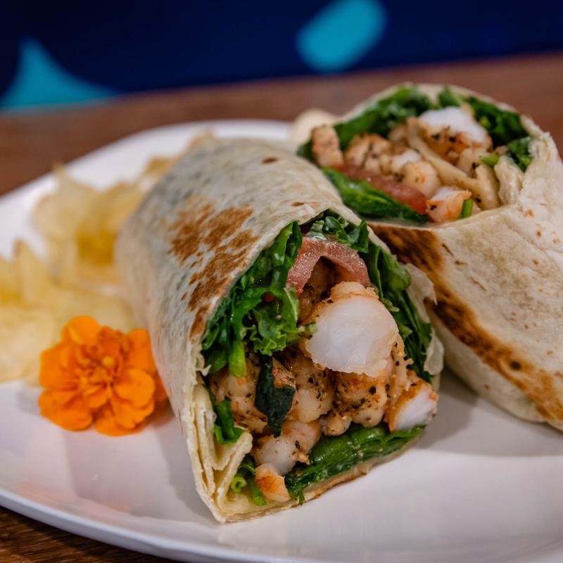 Close-up of a seafood burrito cut in half, on a white plate with potato chips and an orange flower garnish.