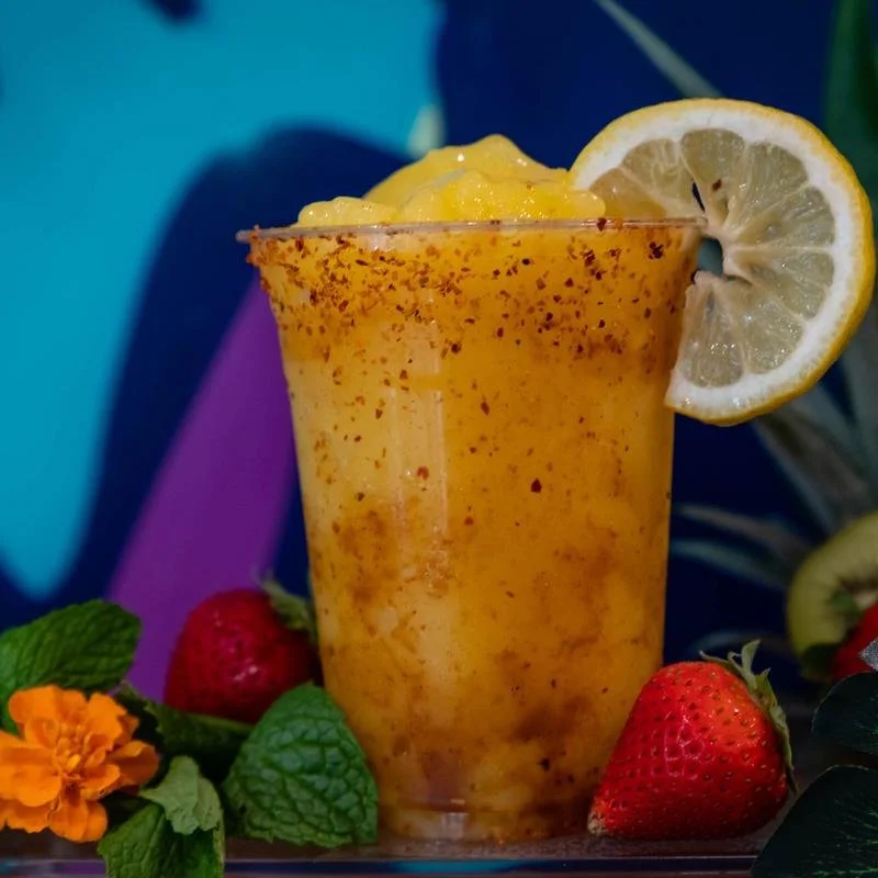 A glass of orange smoothie garnished with a lemon slice, surrounded by strawberries, mint leaves, and an orange flower, against a colorful background.