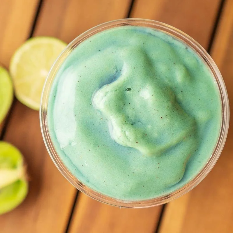 A cold, blended, green-colored beverage in a clear plastic cup with a lid, on a wooden surface with lime slices nearby.