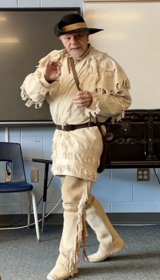 Mike Guli dressed in a Rifleman costume, including a wide-brimmed hat, fringed shirt, buckskin pants, and moccasins, standing in a classroom setting.