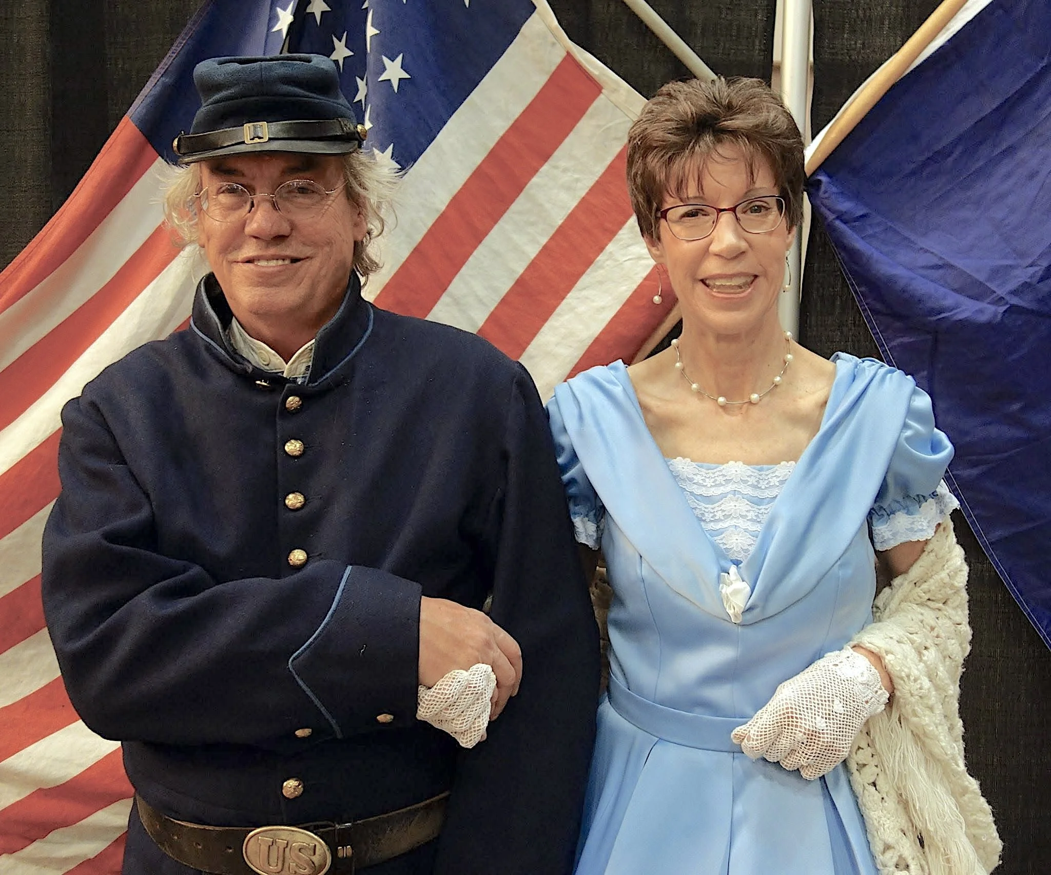 A man and woman dressed in historical costumes, standing in front of American flags at an event or celebration.