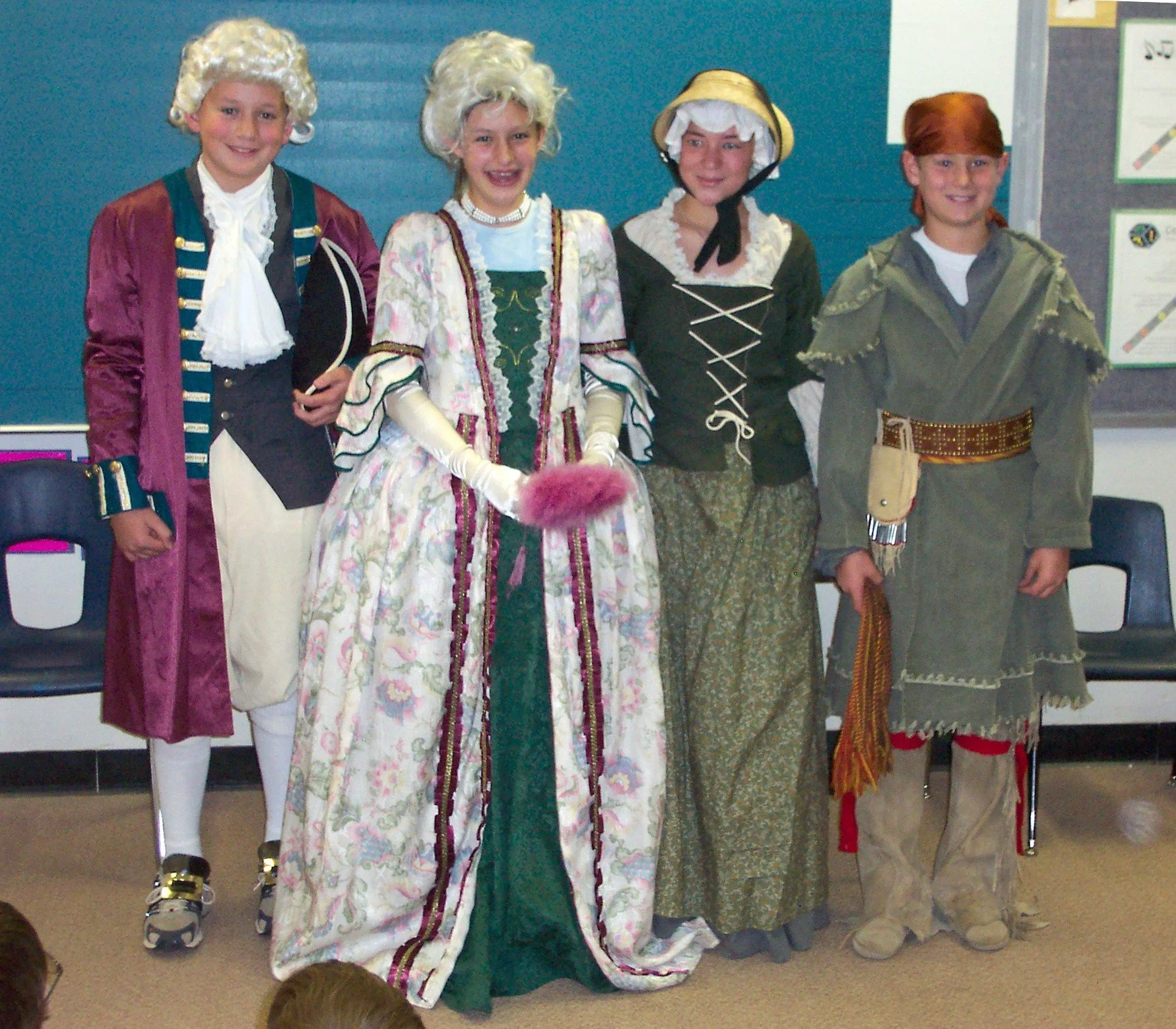 Group of children dressed in 18th-century costumes, including wigs and period attire, standing indoors in front of a blue wall with educational posters.