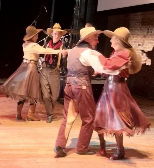 Four people dressed in cowboy attire dancing on stage, with two men and two women, all wearing cowboy hats and vintage-style clothing.