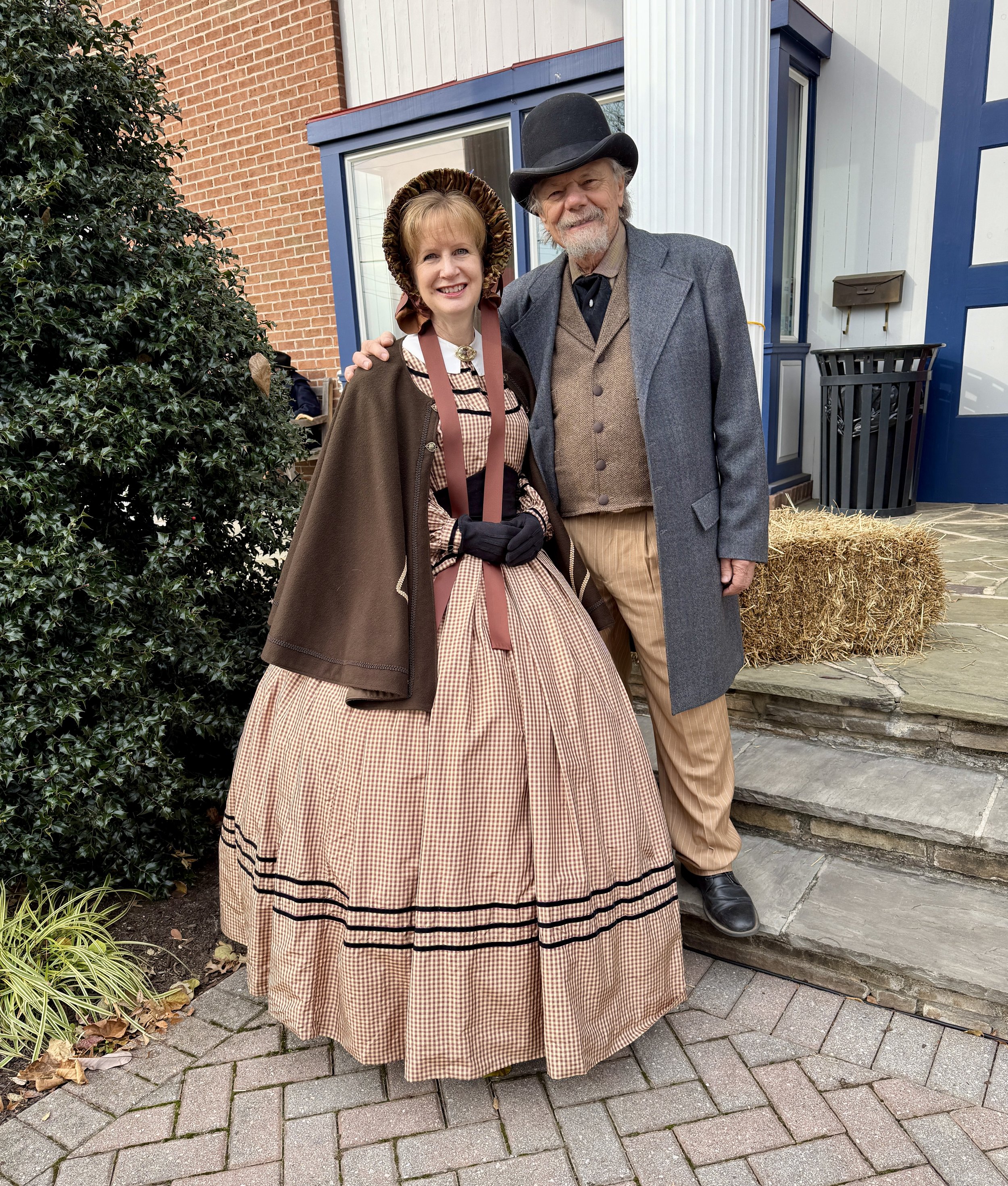 Sharon and Mike Guli in Civil War outfits in front of a building in Gettysburg, PA. She wears a silk gingham dress with black trims, a bonnet, and cape, and he wears a gray frock coat, brown vest, tan pants, and a derby.