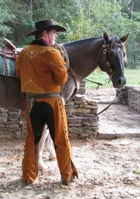 A man dressed in Old West Vaquero attire standing next to a black horse outdoors in a wooded area.