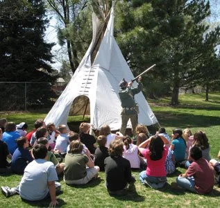 A man giving a presentation or storytelling session to a group of children outdoors in front of a teepee tent.