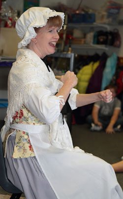 Sharon Guli, in a Revolutionary War/Colonial day costume with white mob cap and apron, sitting and smiling as she teaches a Colonial program to a group of students.