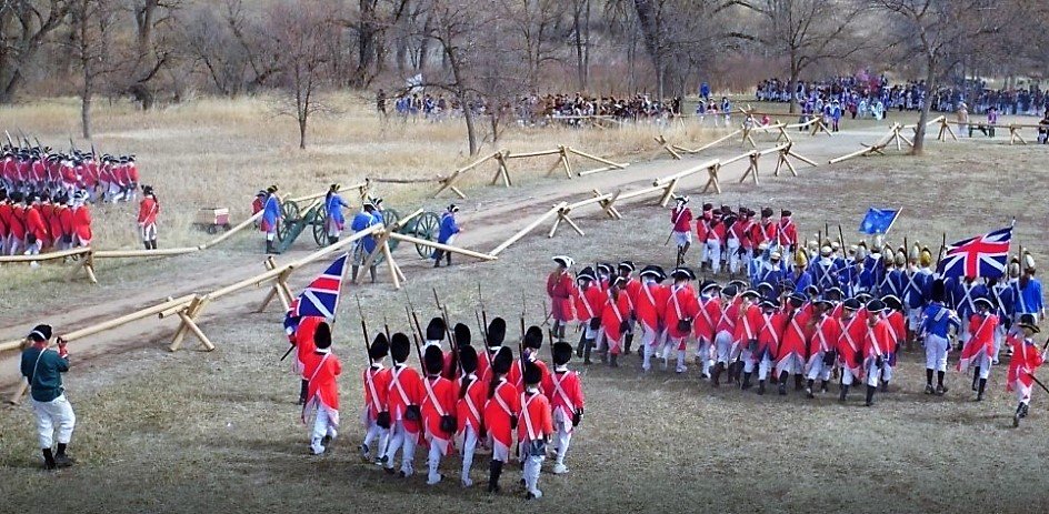 Students at a Revolutionary War Battle Reenactment, marching in uniform across a field, with fencing and cannons along a road.