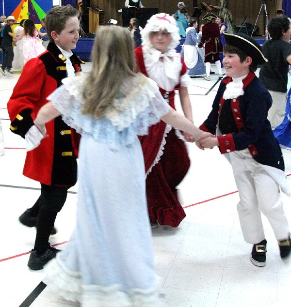 Children in historical costumes dancing in a gym at a Colonial Ball.