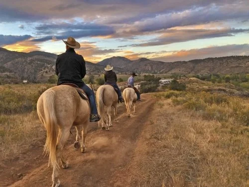 Three people riding horses along a dirt trail in a rural landscape with mountains and a cloudy sunset sky.