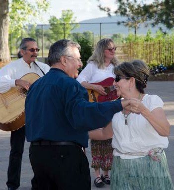 A group of four adults, two men and two women, outdoors enjoying a music-themed event. One man plays guitar, while the others are dancing and smiling.