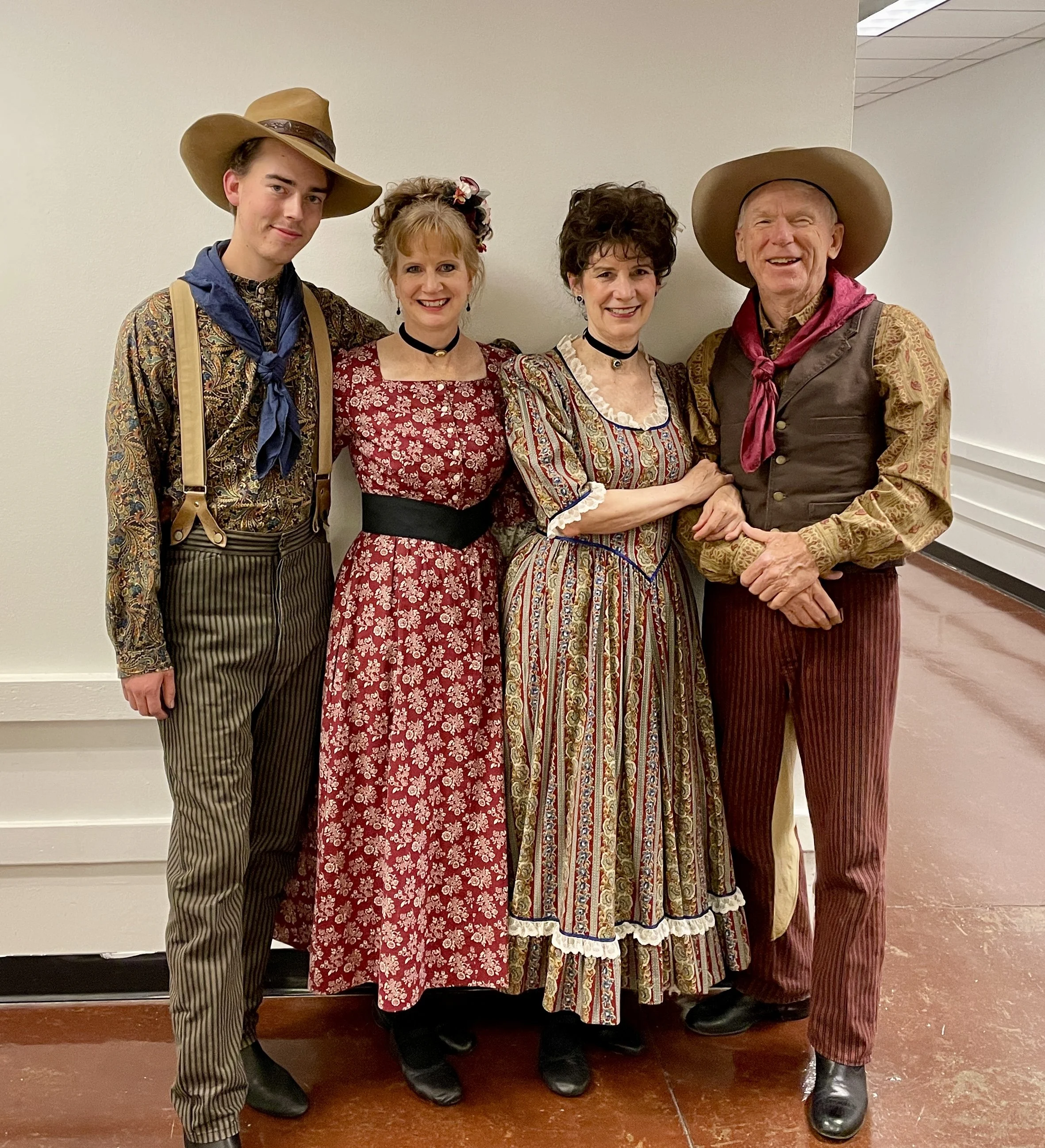 Four people dressed in traditional and historical Western or pioneer costumes, standing together indoors against a plain wall, smiling for the photo.